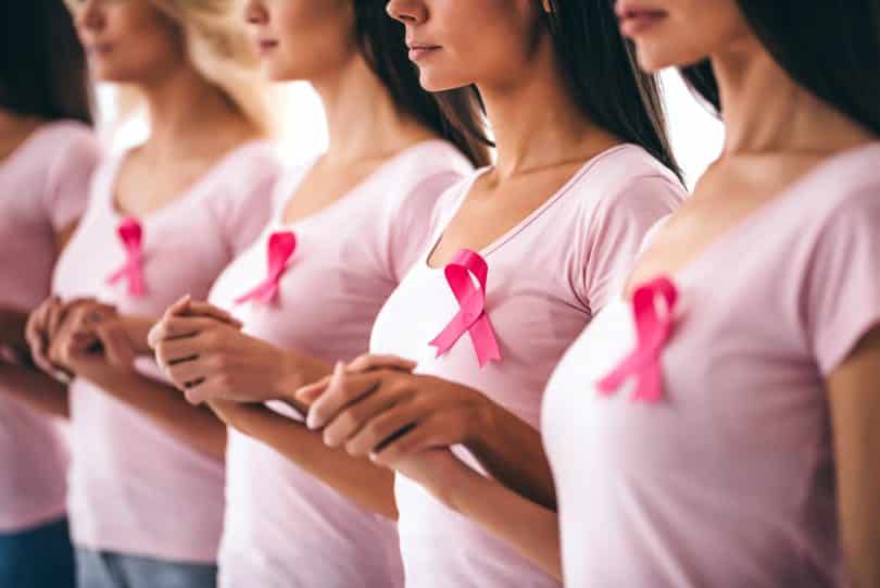 Quatro mulheres vestidas de rosa com as mãos dadas usando fitas da consciência também rosa