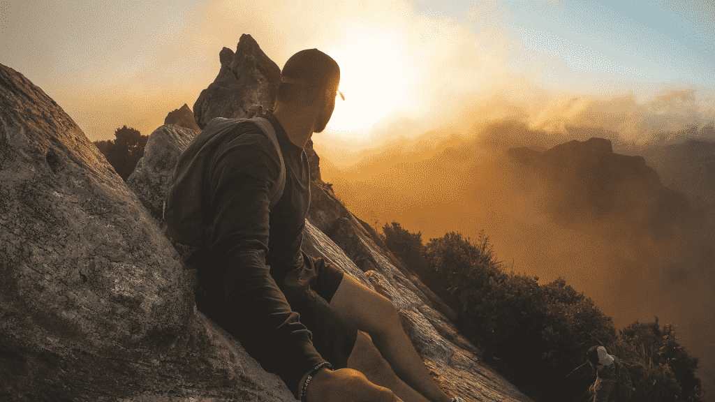 Um homem sentado no cume de uma montanha. Ele contempla o sol.
