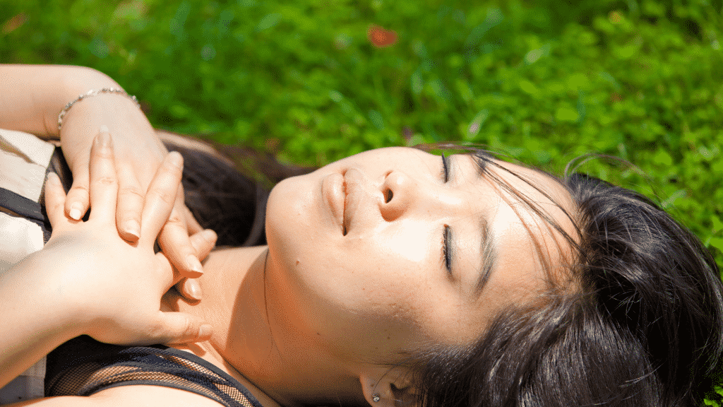 Mulher deitada em um gramado com as mãos no peito e olhos fechados, tomando sol