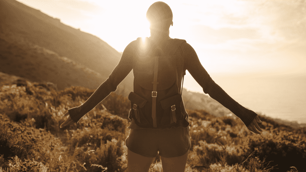 Pessoa de mochila nas costas em uma montanha com os braços abertos, tomando a luz do sol