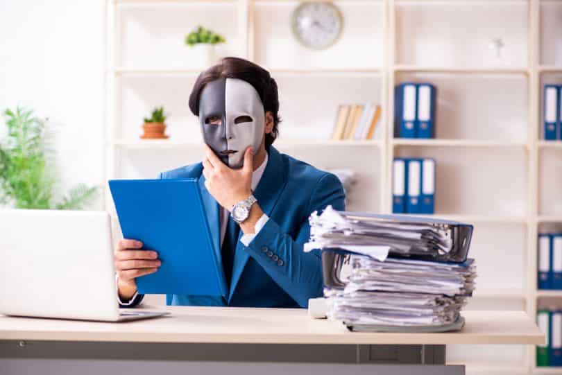 Um homem em frente a um computador. Ele utiliza uma máscara.
