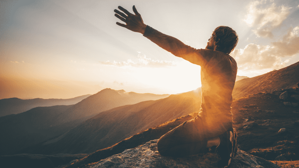 Um homem ajoelhado. Ele ergue seus braços em direção ao céu. Ao fundo, grandes e extensas montanhas esclarecidas pelo sol.