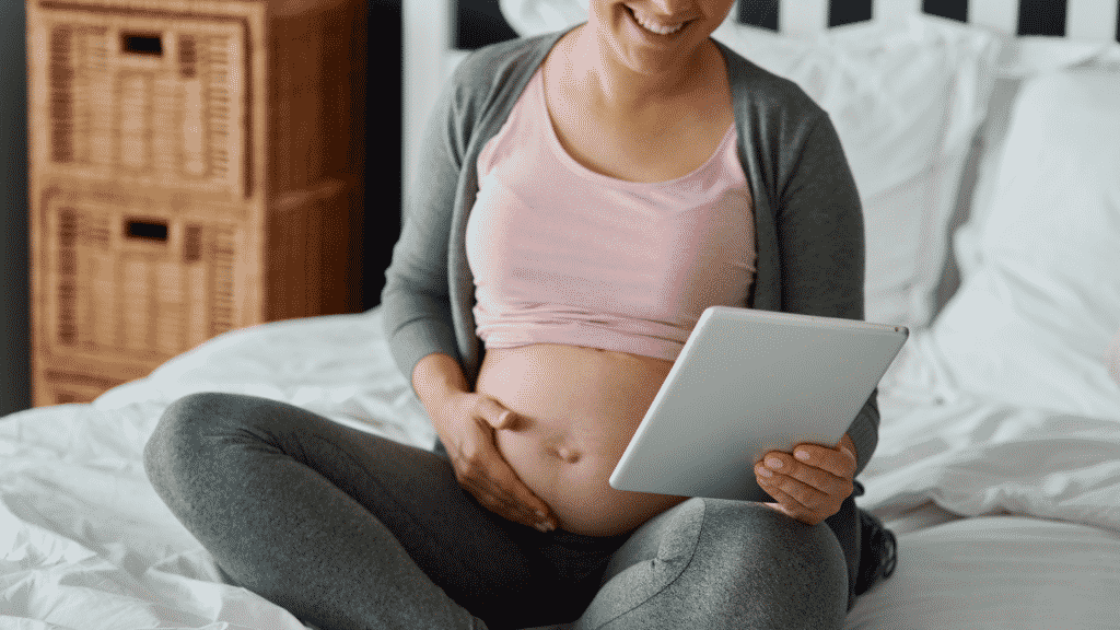 Numa cama, uma mulher grávida sorrindo. Sua mão esquerda acaricia a sua barriga, e a sua mão direita segura um tablet.