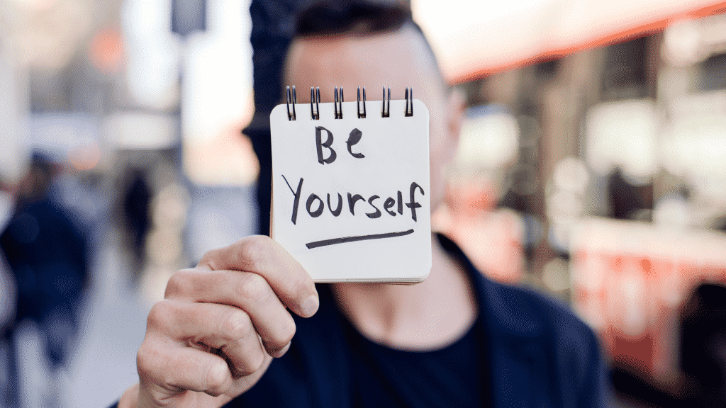 Um homem segurando um pequeno bloco de notas que carrega a frase "be yourself" escrita.
