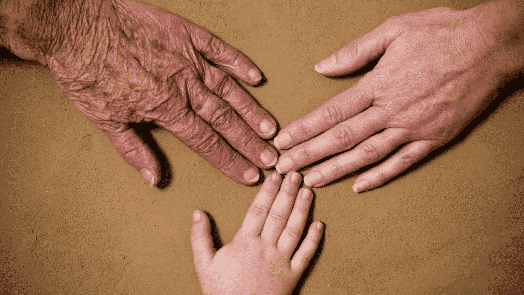 Mãos de diferentes pessoas postas na superfície de uma mesa. À esquerda e em plano fotográfico superior, uma mão envelhecida; à direita, concomitantemente, uma mão de uma mulher adulta. Abaixo de ambas as supracitadas, uma mão de uma criança.