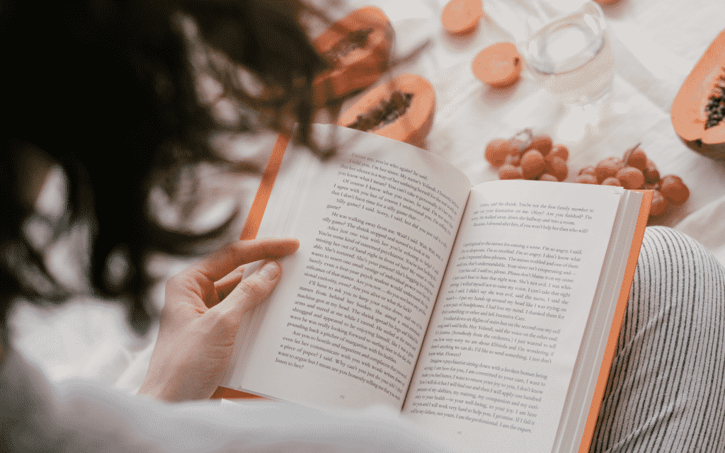 Uma mulher lendo um livro. À frente dela, frutas dispostas numa toalha de mesa.