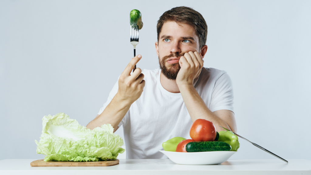 Um homem "garfando" um pepino. Na mesa à qual ele se senta, repolho sobre uma tábua e uma tigela de verduras.