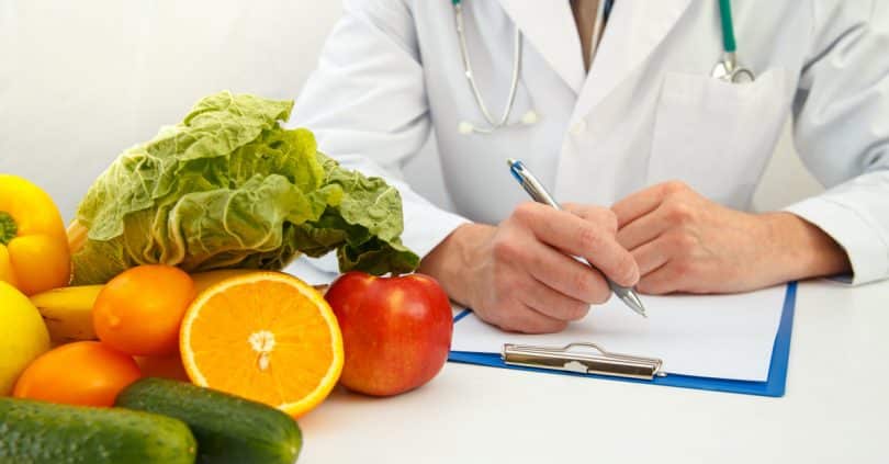 À esquerda, frutas e verduras. À direita, um médico de jaleco escrevendo alguma coisa em seu caderno.