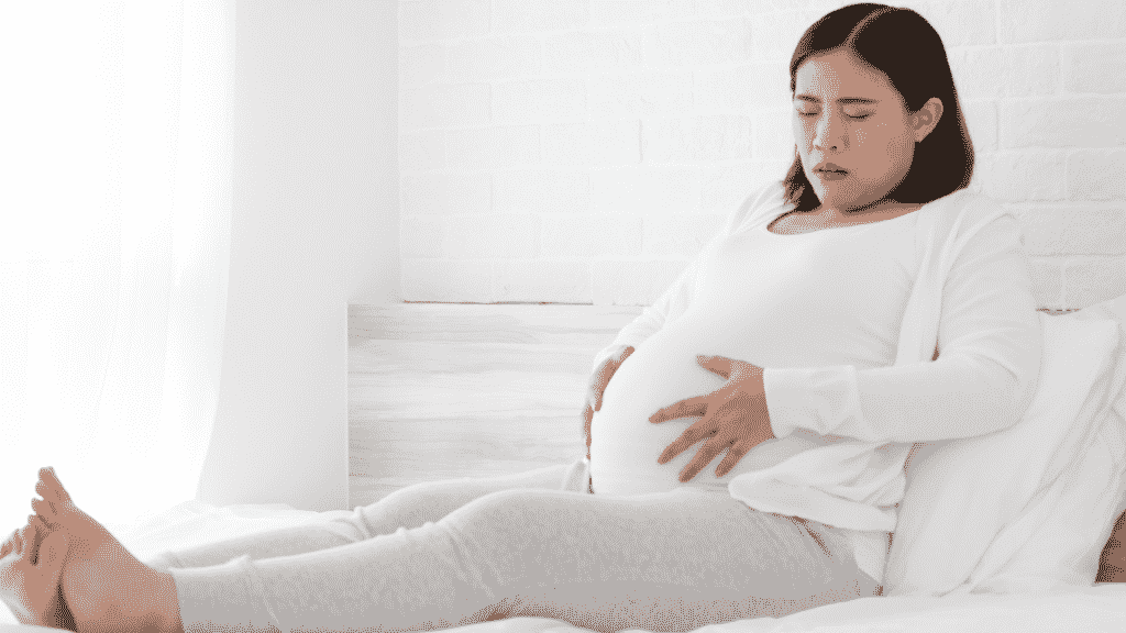 Uma mulher grávida pressionando, numa cama, sua barriga. Ela exibe uma forte expressão de dor.