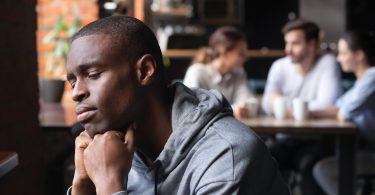 Um homem negro isolado apresentando um semblante de tristeza. Ao fundo, um grupo de pessoas sentados à mesa, conversando e sorrindo.