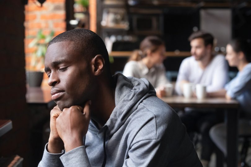 Um homem negro isolado apresentando um semblante de tristeza. Ao fundo, um grupo de pessoas sentados à mesa, conversando e sorrindo.