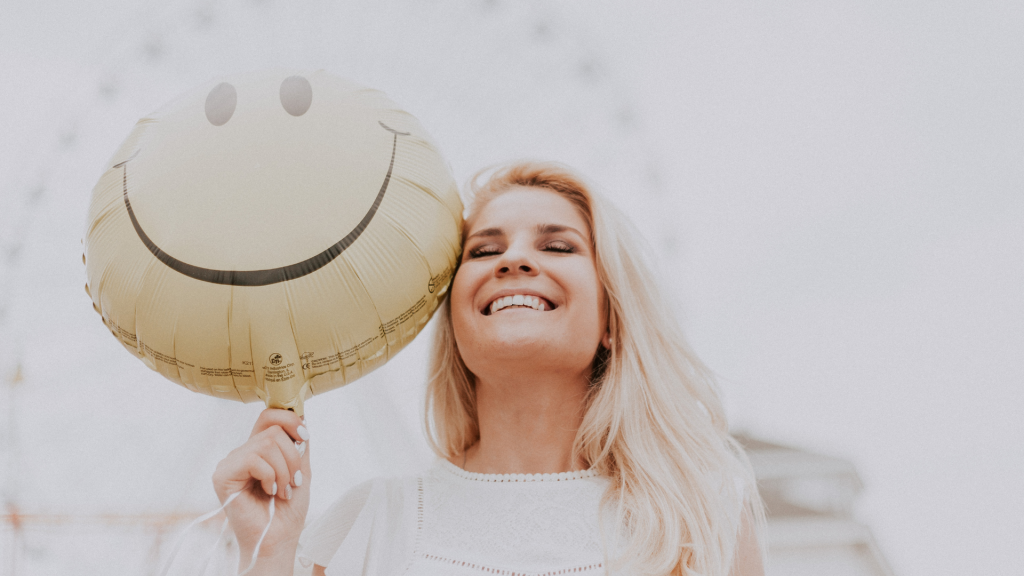 Uma mulher loira sorrindo. Ela segura um balão que tem uma feição sorridente ilustrada no seu centro.