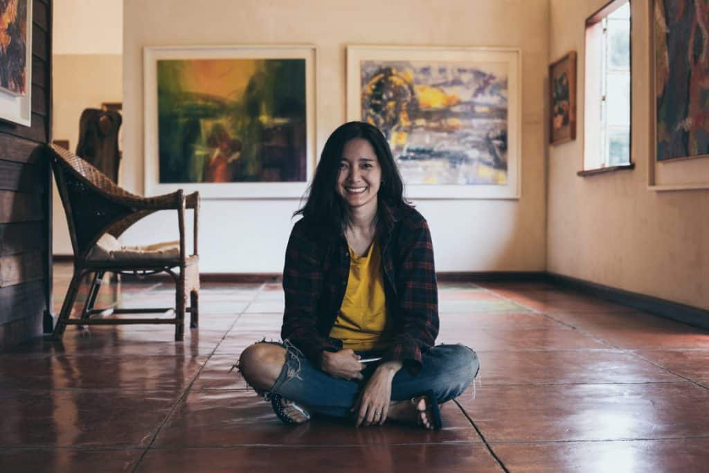 Mulher sentada no chão de uma sala com quadros na parede. Ela está sorrindo.