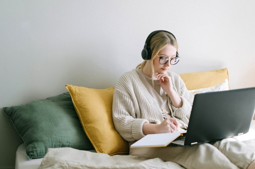 Mulher sentada na cama com o notebook no colo e alguns cadernos de anotação.