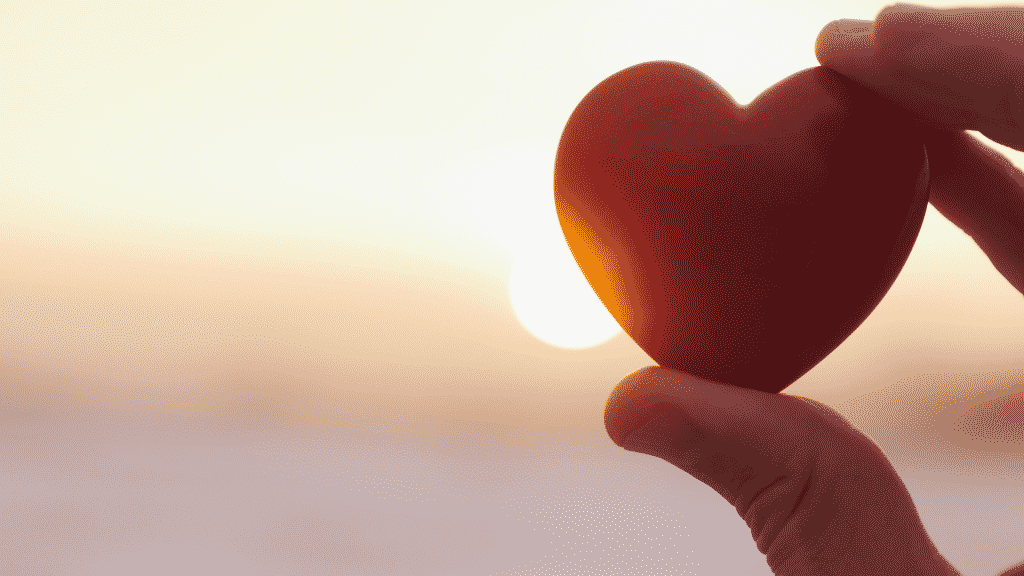 Uma mão segurando uma miniatura de coração em frente a um sol forte.