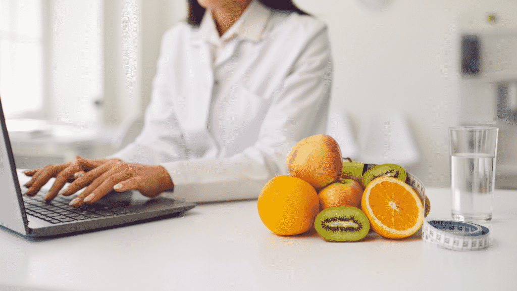 Uma mulher, trajando uma camisa social, utilizando um notebook. À sua direita, frutas, uma fita métrica e um copo d'água.