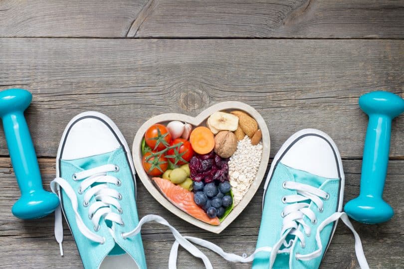 Um par de tênis e, no meio destes, uma vasilha cujo formato é de um coração. Ela comporta diferentes alimentos. Ao lado de cada par de tênis, halteres dispostos em polos/lados opostos.