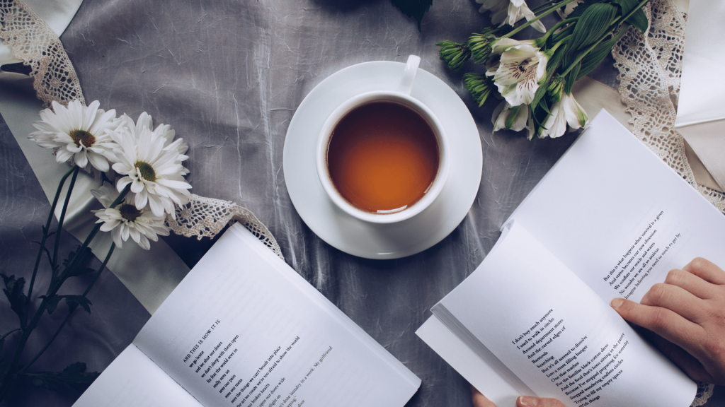 Uma mão segurando um livro de poesia em uma mesa na qual se encontra outro livro, flores e uma xícara de chá