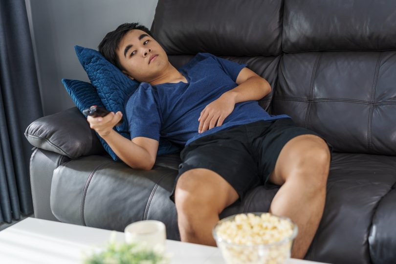 Homem semi-deitado em um sofá com o controle apontado para a televisão.