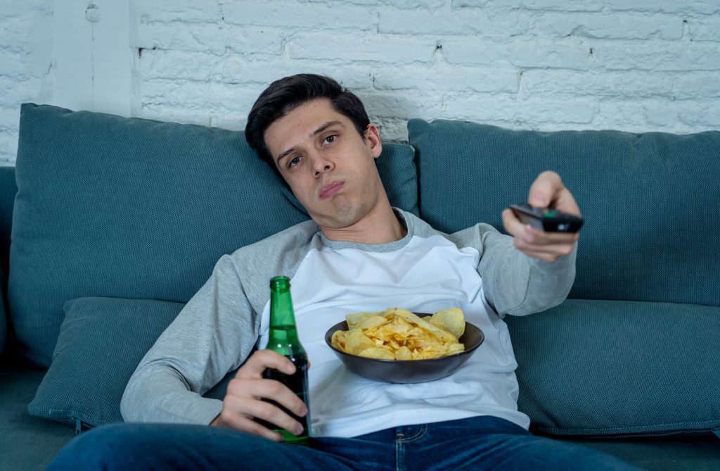 Homem sentado com má postura no sofá segurando uma garrafa de cerveja e um pote de salgadinho.