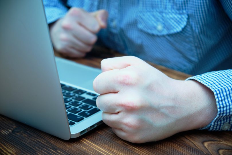 Mão masculina batendo em uma mesa ao lado de um notebook.