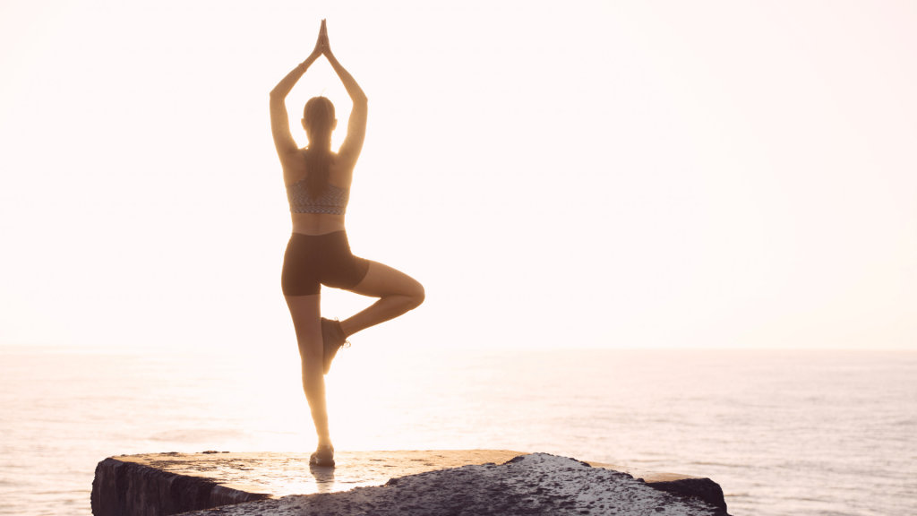 Mulher praticando Yoga em cima de uma pedra com vista para o oceano enquanto o sol está poente