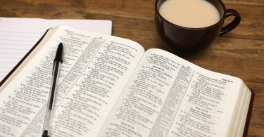 Bíblia aberta em uma mesa. Ao lado há uma xícara com café com leite.