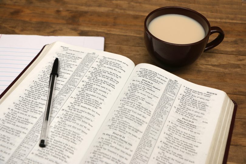 Bíblia aberta em uma mesa. Ao lado há uma xícara com café com leite.