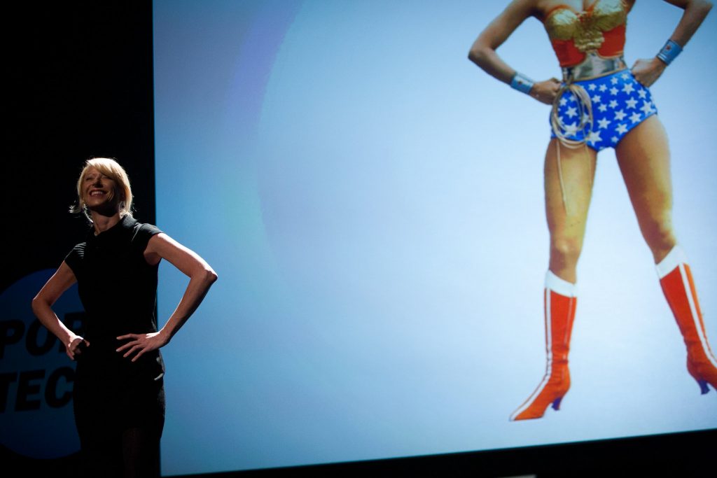 Amy Cuddy, uma mulher loira roupa preta fazendo a pose da mulher maravilha em palestra.