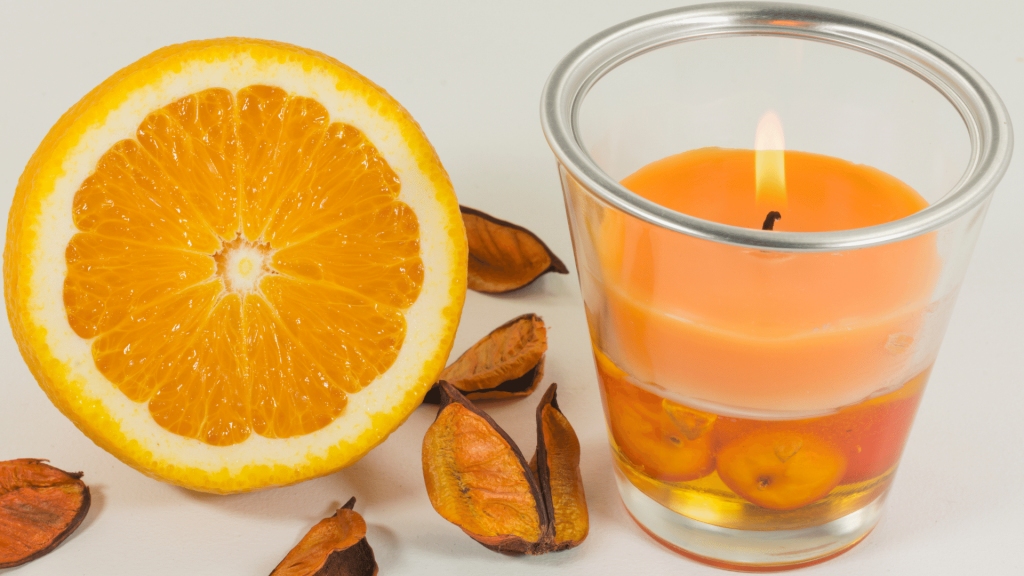 Uma vela aromática de laranja ao lado de uma rodela da fruta em uma superfície branca