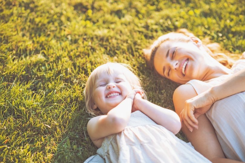 Uma criança deitada sobre a grama. À direita, uma mulher. Ambas sorriem.