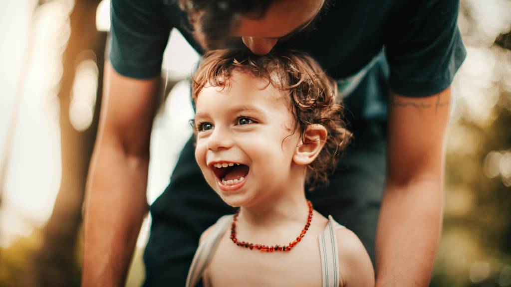 Uma criança sorrindo. Em plano superior a ela, um adulto beijando-a.