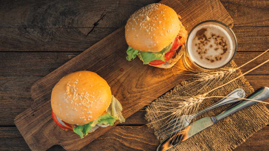 Dois lanches recheados com hambúrguer alface e tomate sobre uma tábua juntamente com uma cerveja apoiada em uma superfície de madeira com um garfo e faca