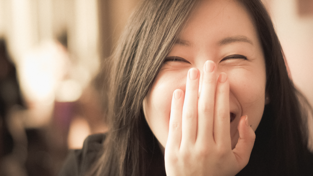 Uma mulher escondendo sua expressão de riso com uma de suas mãos.