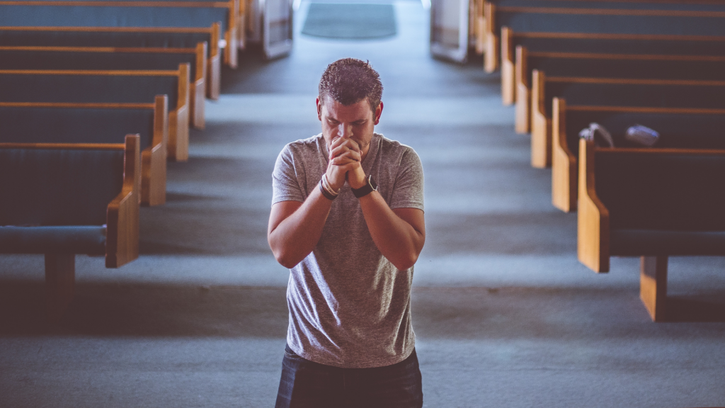 Um homem ajoelhado realizando uma oração. Ao fundo, bancos de igreja.