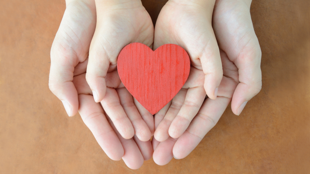 Ao fundo, um par de mãos de um adulto comportando duas mãos de uma criança. Nas palmas das mãos da criança, um coração de madeira.