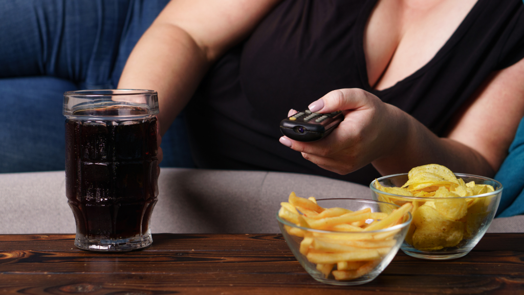 Na superfície de uma mesa, quitutes e salgados. Ao lado destes, um copo que contém refrigerante. Em plano posterior, uma pessoa deita sobre um sofá e segura um controle remoto.