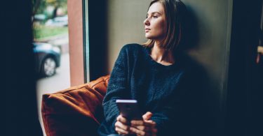 Mulher sentada próxima à janela. Ela está segurando o celular e está olhando para fora.