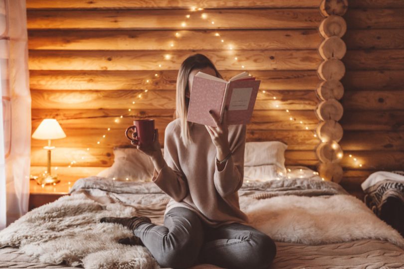 Mulher sentada na cama segurando um livro que está lendo que acaba cobrindo seu rosto e na outra mão uma caneca de café