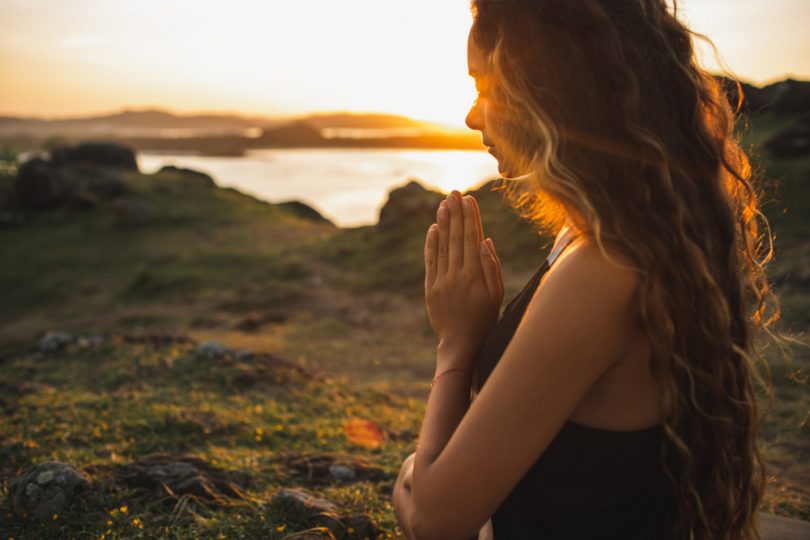 Mulher com as mãos juntas em oração em um lugar iluminado pelo sol da tarde.