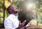 Homem negro com a cabeça para cima. Ele está sorrindo e as mãos estão com as palmas abertas para cima. Ele está orando.