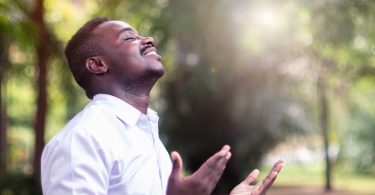 Homem negro com a cabeça para cima. Ele está sorrindo e as mãos estão com as palmas abertas para cima. Ele está orando.