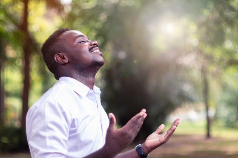 Homem negro com a cabeça para cima. Ele está sorrindo e as mãos estão com as palmas abertas para cima. Ele está orando.