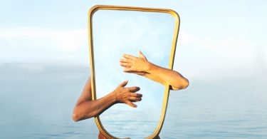 Pessoa branca abraçando espelho com mar ao fundo.