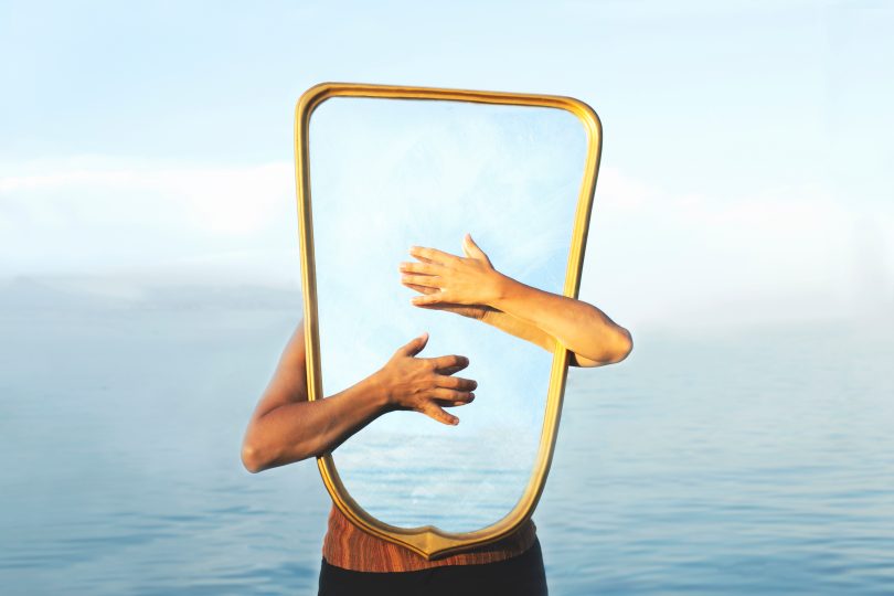 Pessoa branca abraçando espelho com mar ao fundo.