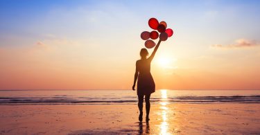 Uma mulher de pé em frente ao mar. Ela segura balões de ar.