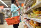 Mulher no mercado segurando um cesto de compras em um de seus braços enquanto mantém seu outro esticado com a mão no queixo analisando as prateleiras