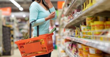 Mulher no mercado segurando um cesto de compras em um de seus braços enquanto mantém seu outro esticado com a mão no queixo analisando as prateleiras