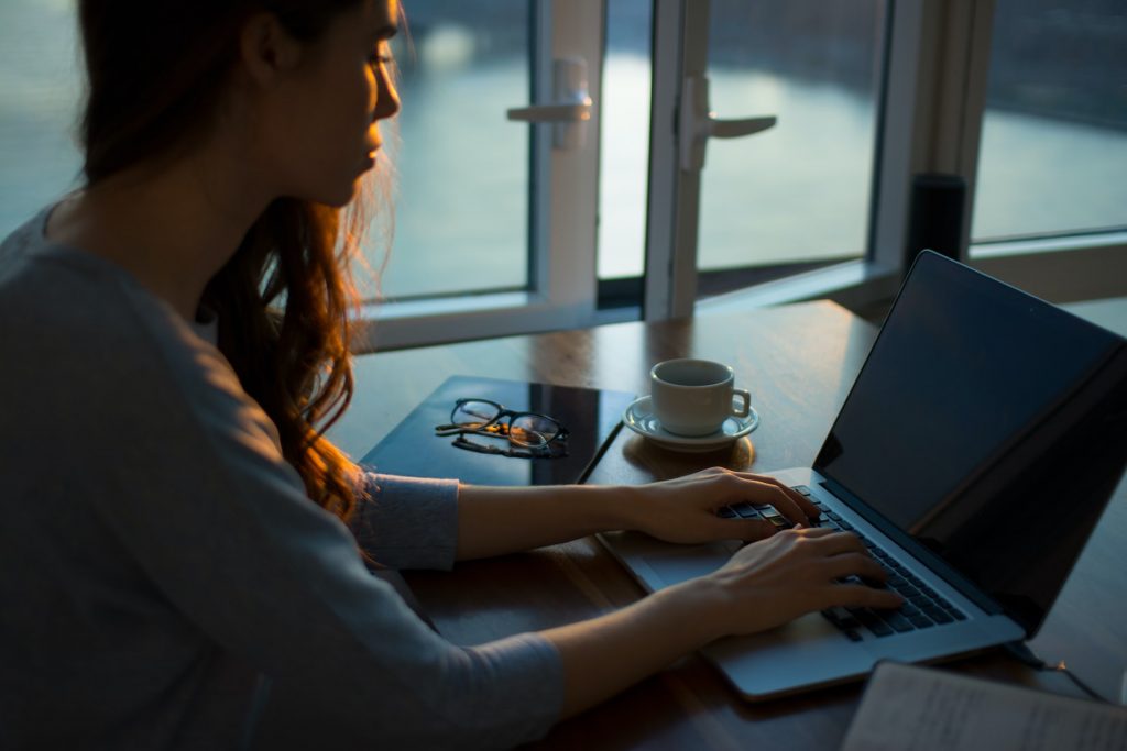 Mulher vestindo roupas cinzas. Ela está sentada em uma mesa mexendo em seu computador.