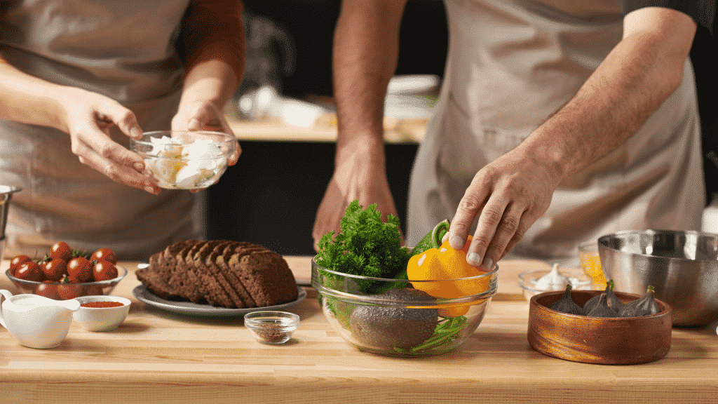 Casal preparando uma refeição, um está segurando o pote de manteiga em ambas as mãos e o outro pegando um pimentão amarelo de um recipiente de vidro